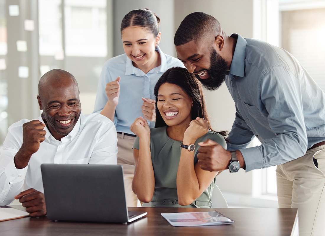 Insurance Solutions - A Team of Employees Celebrating a Good Job While Sitting and Standing in Front of a Laptop at a Wooden Table